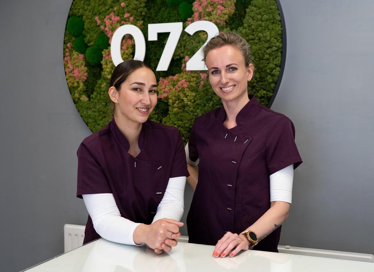Två kvinnor i matchande plommonfärgade uniformer står bakom ett vitt skrivbord och ler. En grön och rosa växtbeklädd väggdekoration med numret "072" syns bakom dem mot en grå vägg.