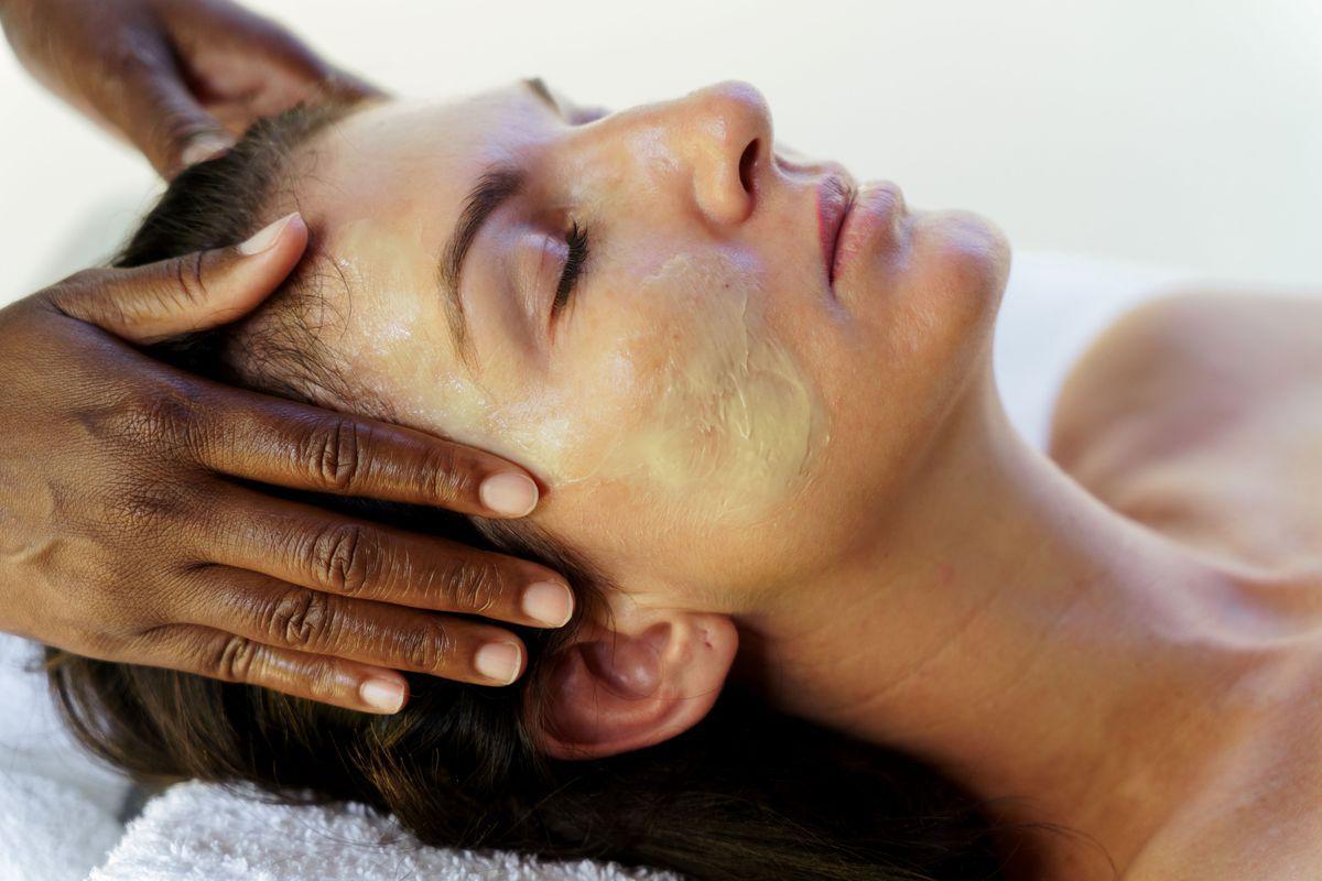 En person med slutna ögon får en avslappnande ansiktsmassage, där händerna försiktigt masserar pannan. Personen har en lätt ansiktsmask på sig och ligger på en vit handduk.