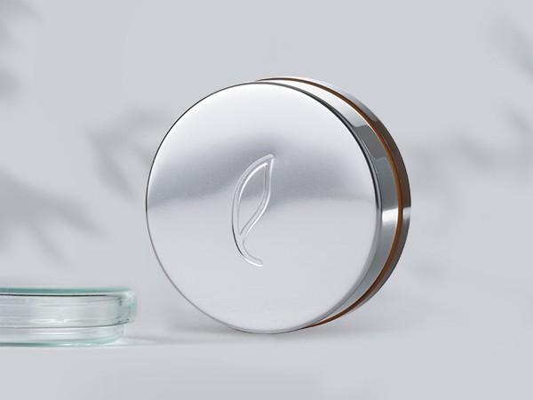 En rund kosmetikbehållare i silver med en enkel bladdesign präglad på locket, mot en ljusgrå bakgrund med en glaskant synlig i förgrunden.