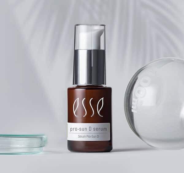 En brun flaska märkt "esse pro-sun D serum" med en silverfärgad pump står på en ljusgrå yta, bredvid ett runt klart föremål och en grund glasskål. Mjuka lövskuggor syns i bakgrunden.