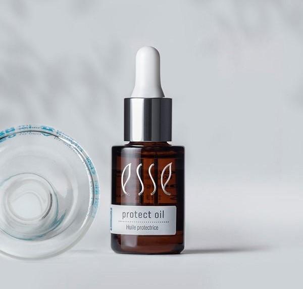 En droppflaska i brunt glas märkt "Esse Protect Oil" står upprätt bredvid ett klart, runt glasföremål på en ljusgrå yta med en enfärgad grå bakgrund.